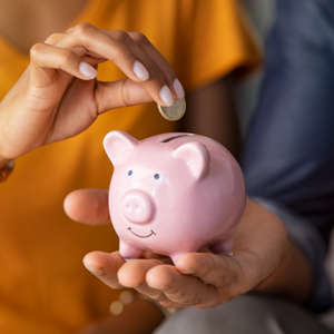 Bild vergr&ouml;&szlig;ern: In einer Hand wird ein rosafarbenes Sparschwein gehalten, eine zweite Person mit gelben T-Shirt wirft Geld in das Sparschwein.