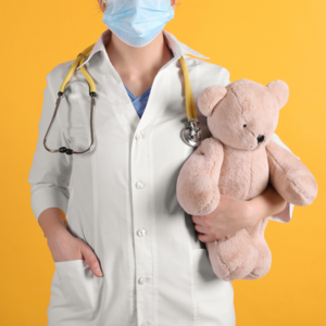 Bild vergr&ouml;&szlig;ern: Abgebildet ist eine &Auml;rztin mit Kittel und Stethoskop vor gelbem Hintergrund, die einen Teddy im Arm h&auml;lt. 